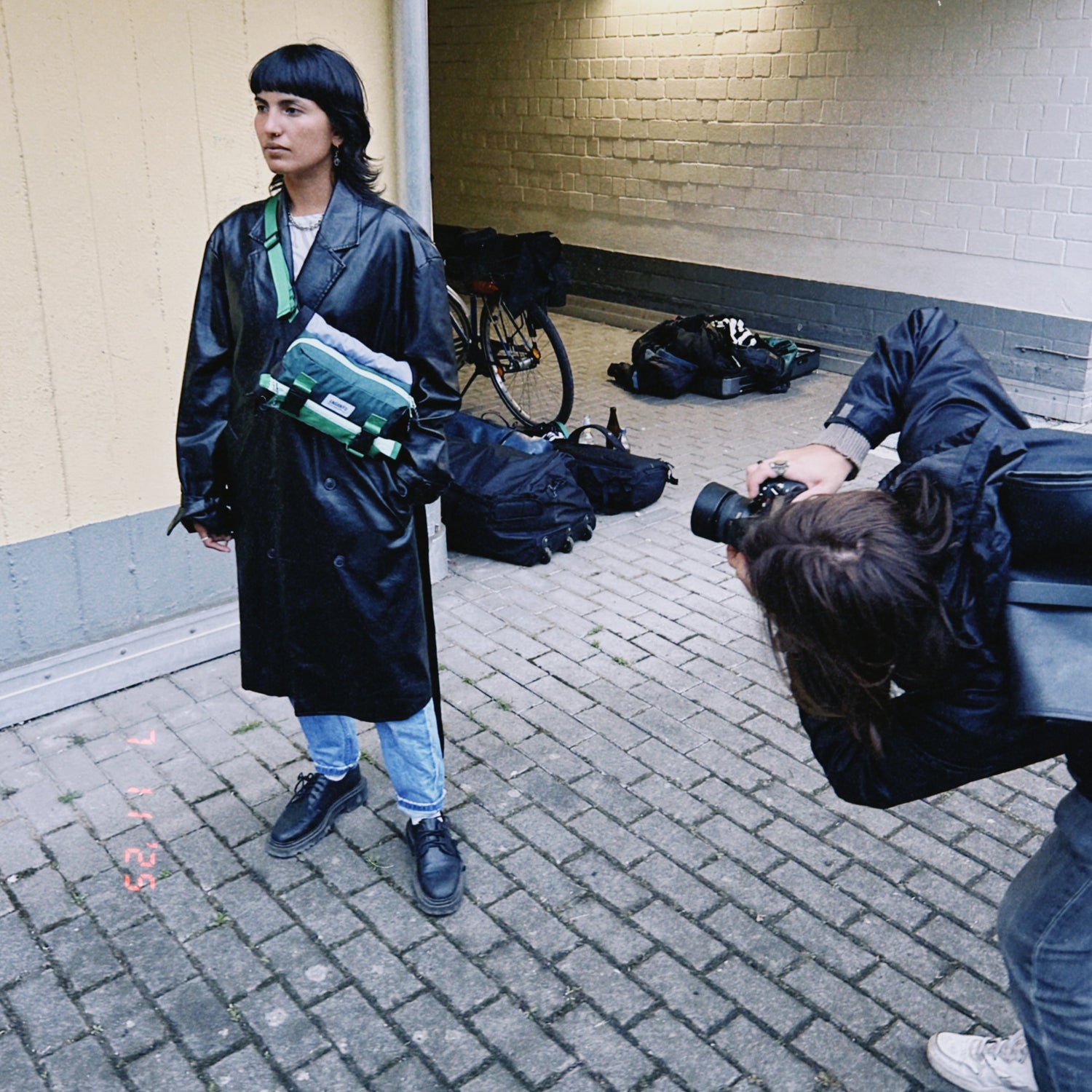 Person in a black raincoat holding a green object, standing next to another person with a camera in an urban setting.