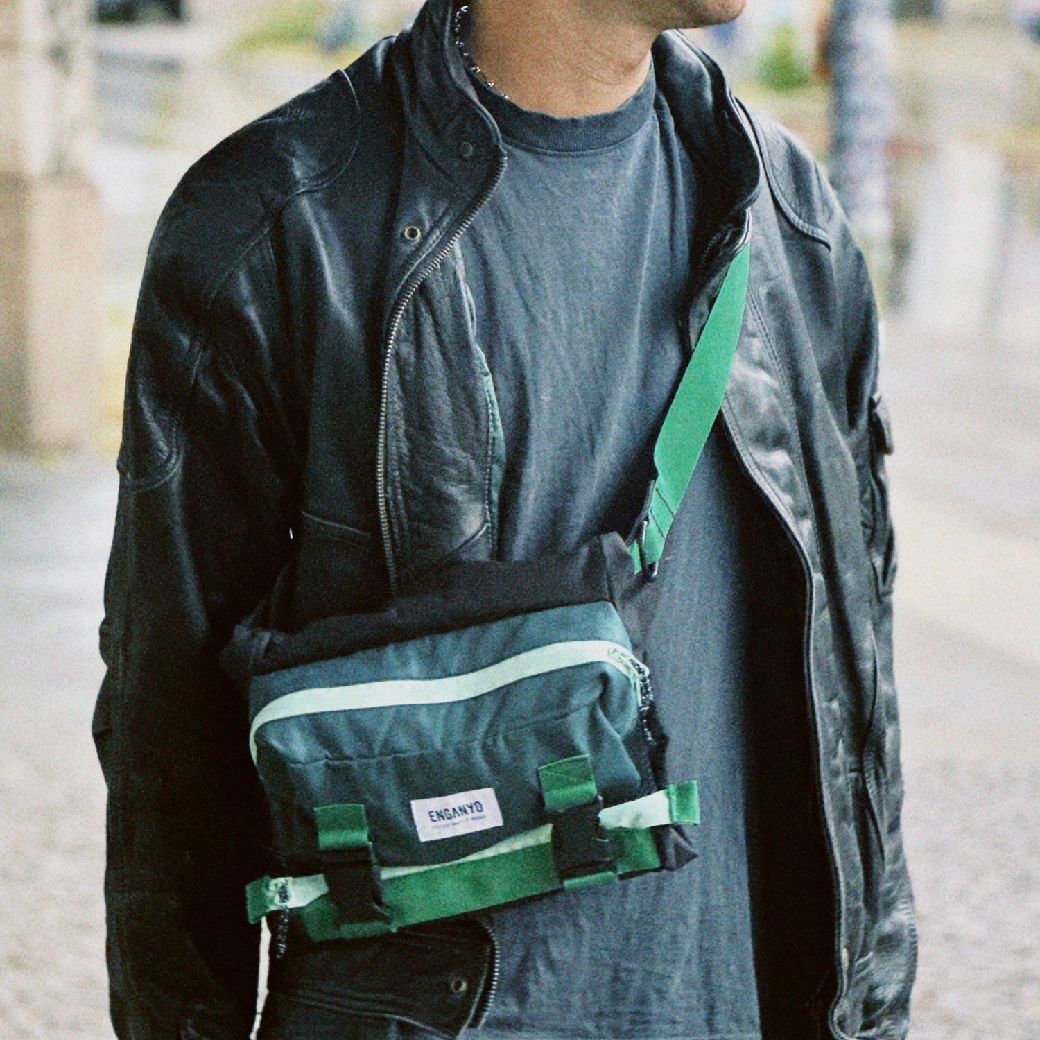 Man wearing a black jacket and cap, carrying a green and black bag on a city street.